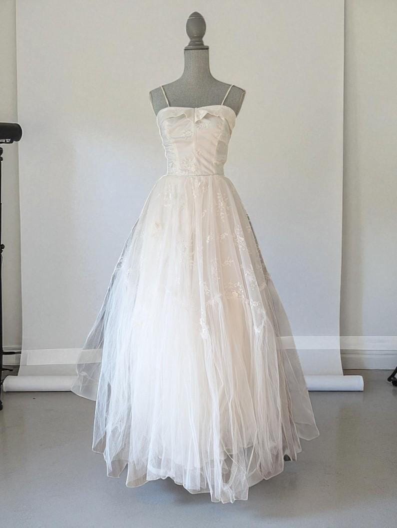 May include: An ivory vintage wedding dress on a mannequin. The dress has a sweetheart neckline, spaghetti straps, and a bow. The fitted bodice leads to a full skirt made of layered tulle, creating a classic silhouette.