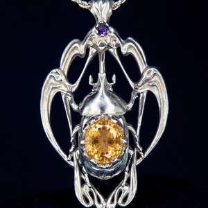 May include: A silver pendant necklace featuring a detailed beetle design with a large, yellow gemstone. The pendant is accented with a smaller, purple gemstone at the top.