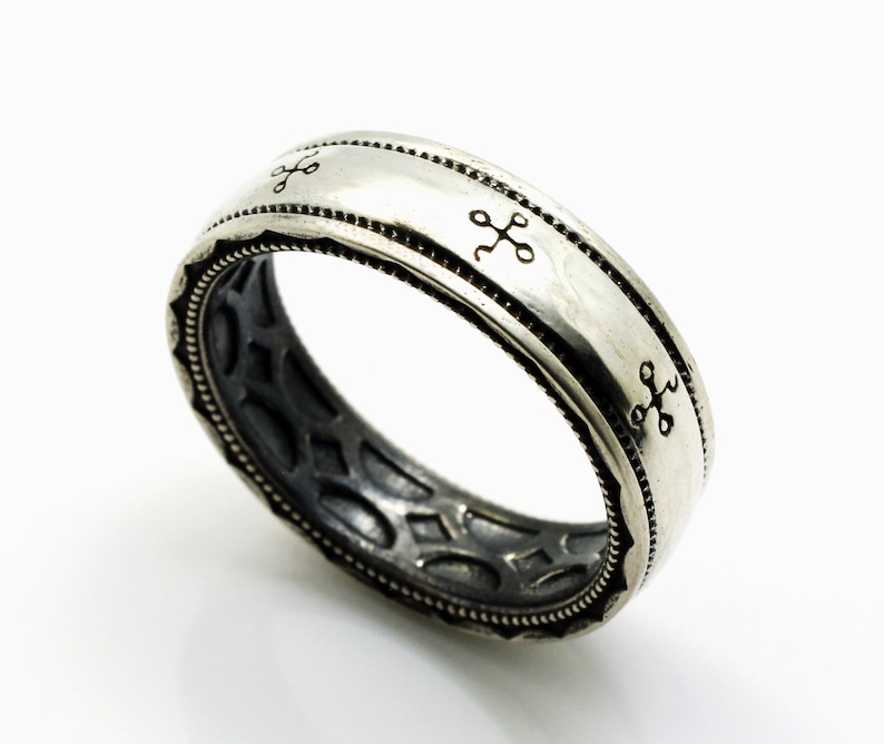 K&ouml;nnte beinhalten: Ein silberner Ring mit detailliertem Design. Der Ring hat ein Band mit kleinen kreuzartigen Symbolen und einem strukturierten Rand. Der innere Teil des Rings hat ein durchbrochenes Muster. Der Ring ist etwa 0,8 cm breit.