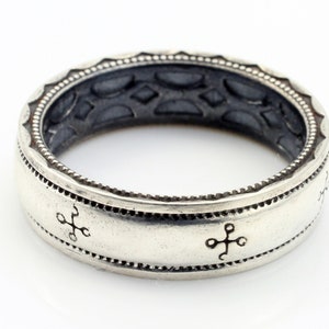 K&ouml;nnte beinhalten: Ein silberfarbener Ring mit einem eingravierten Design. Der Ring hat ein Band mit kreuzartigen Symbolen und einem detaillierten Rand. Der innere Teil des Rings hat ein strukturiertes Muster. Ein klassisches Schmuckst&uuml;ck.