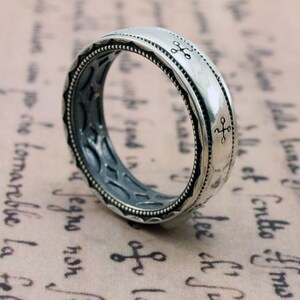 K&ouml;nnte beinhalten: Ein silberner Ring mit aufwendigem Design. Der Ring hat ein detailliertes Muster an den R&auml;ndern und kleine kreuzartige Symbole. Das Innere des Rings hat ein strukturiertes, dunkelgraues Aussehen.