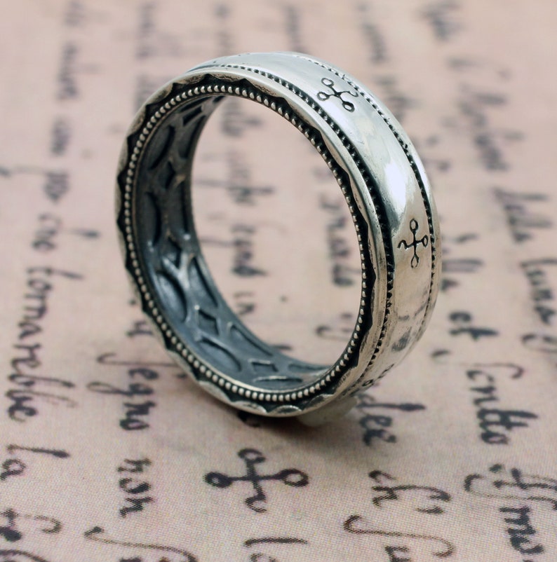 K&ouml;nnte beinhalten: Ein silberner Ring mit aufwendigem Design. Der Ring hat ein detailliertes Muster an den R&auml;ndern und kleine kreuzartige Symbole. Das Innere des Rings hat ein strukturiertes, dunkelgraues Aussehen.