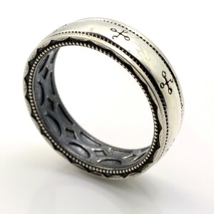 K&ouml;nnte beinhalten: Ein silberfarbener Ring mit aufwendigem Design. Der Ring hat eingravierte kreuzartige Symbole auf dem Band und einen detaillierten Rand. Die Innenseite des Rings hat ein dunkles, strukturiertes Muster. Der Ring wird vor wei&szlig;em Hintergrund gezeigt.