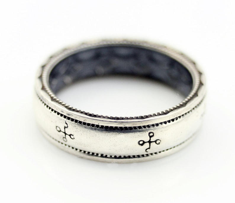 K&ouml;nnte beinhalten: Ein silberner Ring mit detailliertem Design. Der Ring hat ein Band mit einer Reihe kleiner, gleichm&auml;&szlig;ig verteilter Punkte und kreuzartigen Symbolen. Der obere Rand hat ein dekoratives Muster. Der Ring ist wahrscheinlich aus Sterlingsilber.