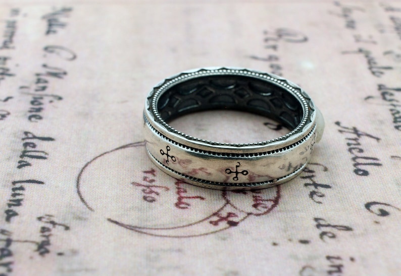 K&ouml;nnte beinhalten: Ein silberner Ring mit detailliertem Design. Der Ring hat ein Band mit kleinen kreuzartigen Symbolen und einem strukturierten Rand. Der Hintergrund ist ein Papier im Vintage-Stil mit verblasstem Text und einem kreisf&ouml;rmigen Stempel.