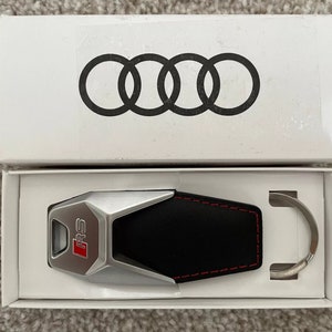 May include: A silver and black key chain with a leather loop and a silver key fob with the letters "RS" in red. The key chain is in a white box with the Audi logo.