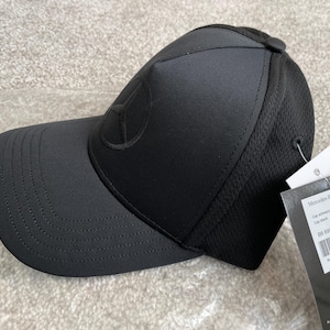 Puede incluir: Gorra de béisbol negra con un logotipo de Mercedes-Benz bordado en la parte delantera. La gorra tiene una parte trasera de malla y una correa ajustable negra.