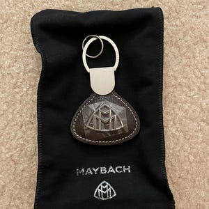 May include: A brown leather keychain with a silver Maybach logo and a silver key ring. The keychain is in a black velvet pouch with the Maybach logo printed on it.