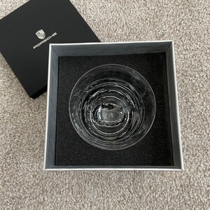May include: A clear, patterned glass tumbler inside a gray box with black foam padding. The box is open, with a black lid featuring the Porsche logo. The tumbler has a circular design and is likely for beverages.