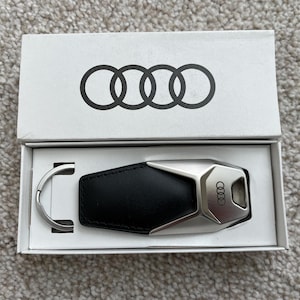 Pode incluir: Chave do carro Audi numa caixa branca. A tampa da caixa apresenta o logótipo da Audi, quatro anéis interligados. A chave tem uma secção em couro preto e uma secção em metal prateado com o logótipo da Audi. Um porta-chaves prateado também está incluído.