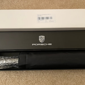 May include: A black umbrella with a silver Porsche logo on a black box with the word "Porsche" in silver lettering. The box is inside a white box with a shipping label.