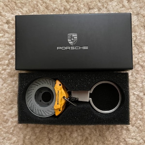 May include: A yellow and gray metal Porsche brake rotor keychain with a silver ring. The keychain is in a black box with the Porsche logo.