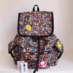 May include: A black backpack with a colorful pattern of video game characters and icons. The backpack has a flap closure, side pockets, and a drawstring closure. A yellow coin is on the flap. The brand name is visible on the side.
