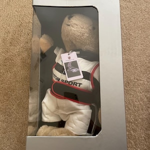 May include: A plush toy bear wearing a white racing suit with black and red accents. The bear is in a clear plastic box with a gray cardboard exterior. The bear has a tag on its chest that says "Porsche Motorsport".