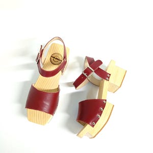May include: A pair of red leather sandals with wooden heels and buckles. The sandals have a small metal logo on the strap.