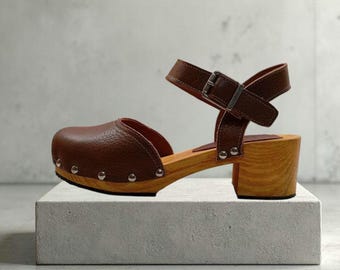 May include: Brown leather clogs with a wooden sole and a block heel. The clogs have a strap with a buckle closure and silver-colored studs along the sole. The heel height is approximately 3 inches.