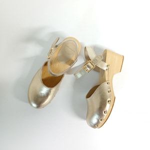 Puede incluir: Un par de zuecos de cuero dorado con tacones de madera y correas de tobillo. Los zuecos tienen un acabado metálico y están adornados con pequeños tachuelas doradas.