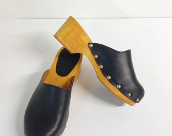 Op de afbeelding: Een paar zwarte leren klompen met houten hakken en gouden studs. De klompen staan op een wit oppervlak.