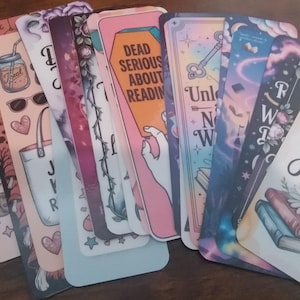 May include: A collection of colorful bookmarks with various designs and text. The bookmarks feature illustrations of books, flowers, and quotes like "Reading is my Jam" and "Dead Serious About Reading". The color palette includes pastels and vibrant hues.