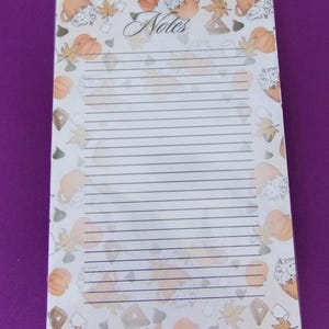 May include: A lined notepad with a decorative border featuring pumpkins, fall leaves, and mugs of coffee. The word "Notes" is written in cursive at the top. The notepad is on a purple surface.