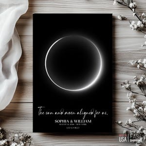 May include: A black and white print featuring a glowing solar eclipse design. The text reads "The sun and moon aligned for us," along with names, a date, location, and zodiac signs. The print is made in the USA.