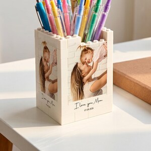 Peut inclure: Un porte-stylo blanc à motif de briques avec une photo personnalisée d'une personne tenant un enfant, portant l'inscription "I love you Mom" et "07.09.2026". Il contient divers stylos colorés et une règle en bois graduée jusqu'à 30 cm.