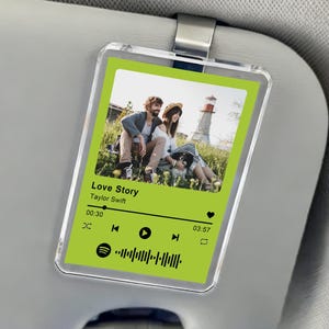 Peut inclure: Un clip de visière de voiture en acrylique transparent avec un design de style Spotify vert et blanc, affichant le texte "Love Story" de Taylor Swift. L'image montre un couple et un phare. Le clip est fixé à une visière de voiture grise.