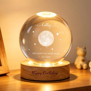 May include: A glowing crystal ball night light with a wooden base. The sphere features a moon and stars design, with the name "Cathy" and the text "THE NIGHT YOU WERE BORN 10.12.2020" engraved. The base reads "Happy Birthday".