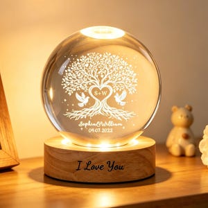 May include: A crystal ball lamp with a tree of life design, two doves, and the text "Sophia & William 04.07.2022". The lamp sits on a wooden base with "I Love You" engraved. The lamp emits a warm, soft light, creating a cosy atmosphere.