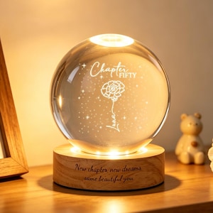 May include: A glowing crystal ball lamp with the words "Chapter Fifty" and "Luna" etched inside, along with a floral design. The lamp sits on a wooden base with the inscription "New chapter, new dreams, same beautiful you."