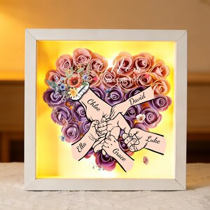May include: A framed, illuminated artwork featuring a heart-shaped arrangement of pink and purple paper roses. The artwork includes illustrations of hands holding each other, with names like "Chloe," "David," "Ella," "Grace," and "Luke" on the hands.