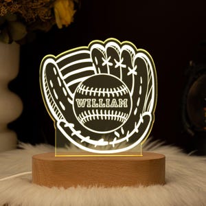 May include: A baseball-themed night light featuring a baseball glove and ball design. The ball has the name "WILLIAM" in the center. The light sits on a wooden base and is illuminated with a soft glow. The overall design is a unique decorative piece.