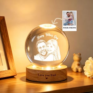 May include: A clear crystal ball with a photo of a man and child, illuminated by a warm light. The base is wooden and inscribed with "Love you Dad." The words "Happy Father's Day" are etched on the ball. A small photo is attached with the words "Your Photo."
