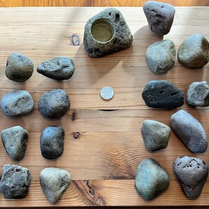 May include: A collection of smooth, gray and green stones arranged on a wooden surface. A small, gray, ceramic candle holder is in the center of the image.