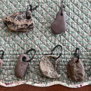 May include: A collection of eight unique rock keychains, each featuring a different shape and texture. The rocks are attached to silver key rings and are displayed on a green and white patterned fabric.