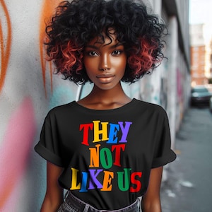 Op de afbeelding: Zwart T-shirt met de tekst "THEY NOT LIKE US" in regenboogkleuren.