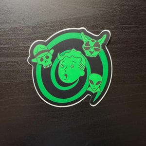 May include: A die-cut sticker featuring a bright green spiral design on a black background. The graphic includes a central cartoon face, a skull wearing a fedora, a cat wearing sunglasses, and an alien head, all rendered in a vibrant green outline.