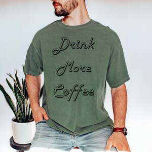 Può includere: Una maglietta verde con il testo "Drink More Coffee" in un font stilizzato.