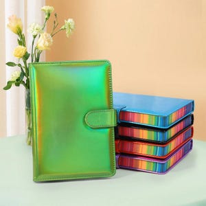 May include: A green iridescent planner with a snap closure is next to a stack of rainbow-edged planners. The planners are on a light green surface, with a vase of flowers in the background.