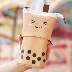 Op de afbeelding: Een beige etui in de vorm van een bubble tea-beker. De etui heeft een lachend gezicht, roze blushvlekken en donkerbruine boba-parels. Het heeft een witte bovenkant en een bruine ritstrekker.