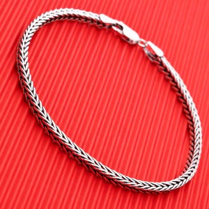 May include: A silver chain bracelet with a braided design. The bracelet is on a red background.