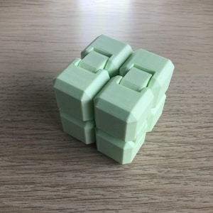 May include: A light green infinity cube fidget toy. The cube is made of several connected blocks, each with a geometric shape. The toy is designed to be manipulated and folded in various ways. The background is a light brown wooden surface.