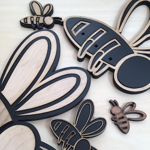 May include: Wooden bee cutouts in various sizes. The bees have a layered design with a natural wood grain and black accents. The wings and bodies are detailed, creating a 3D effect. These decorative pieces are ideal for craft projects.