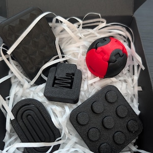 May include: A collection of black and red 3D printed fidget toys including a rectangular textured card, a small square block with nine circular protrusions, a black oval piece with concentric ridges, and a red and black spherical puzzle toy.