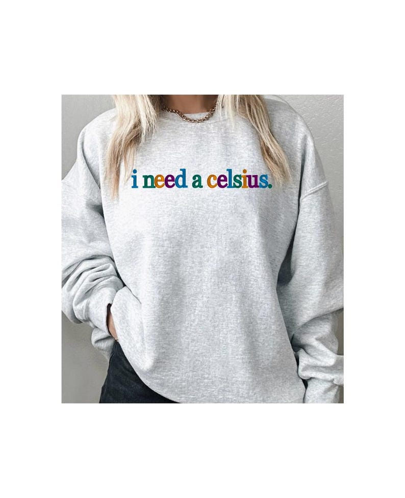 May include: A light gray sweatshirt with the phrase "i need a celsius" printed across the chest in rainbow-colored letters. The sweatshirt has long sleeves and a crew neck.
