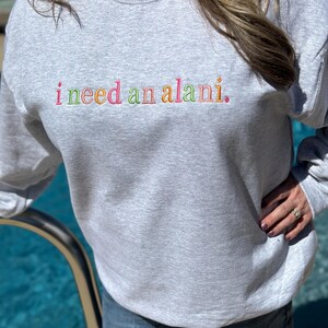 May include: A light gray sweatshirt with the embroidered text "i need an alani." The text is in a rainbow of colors. The sweatshirt is a crew neck style and is being worn with blue jeans.