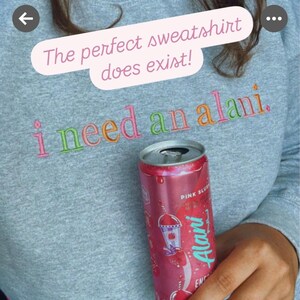 May include: A gray sweatshirt with the embroidered phrase "i need an alani." A pink can of Alani Energy drink is held in front of the sweatshirt. The text "The perfect sweatshirt does exist!" is above the embroidered text.