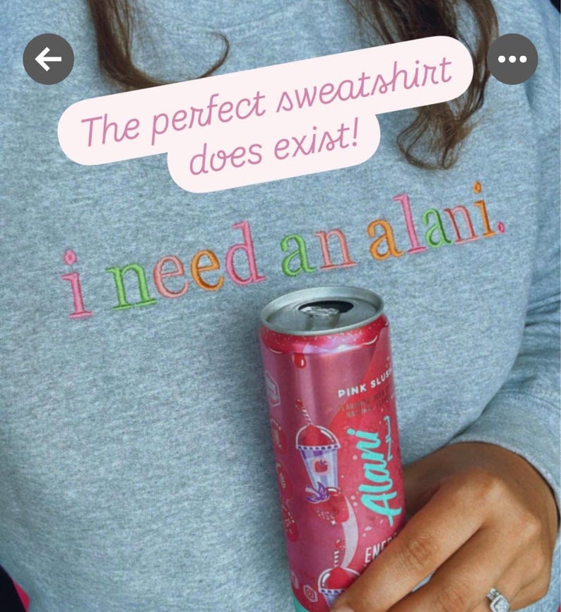May include: A gray sweatshirt with the embroidered phrase "i need an alani." A pink can of Alani Energy drink is held in front of the sweatshirt. The text "The perfect sweatshirt does exist!" is above the embroidered text.