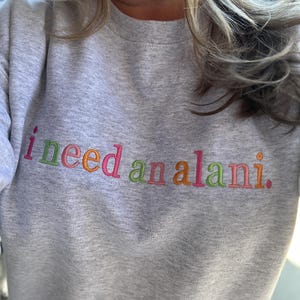 May include: A light gray sweatshirt with the embroidered text "i need an alani" in colorful thread. The text is in a playful, multi-colored font, with each word in a different color.