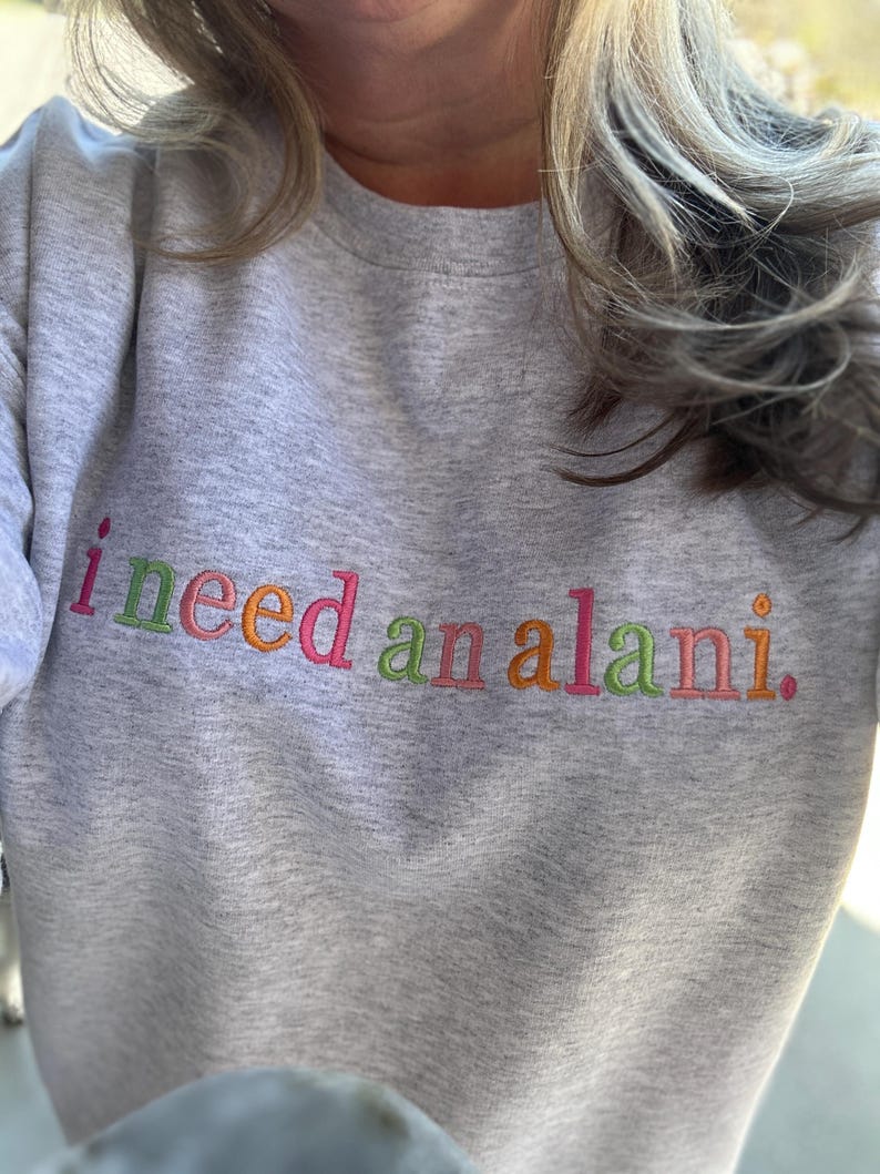 May include: A light gray sweatshirt with the embroidered text "i need an alani" in colorful thread. The text is in a playful, multi-colored font, with each word in a different color.